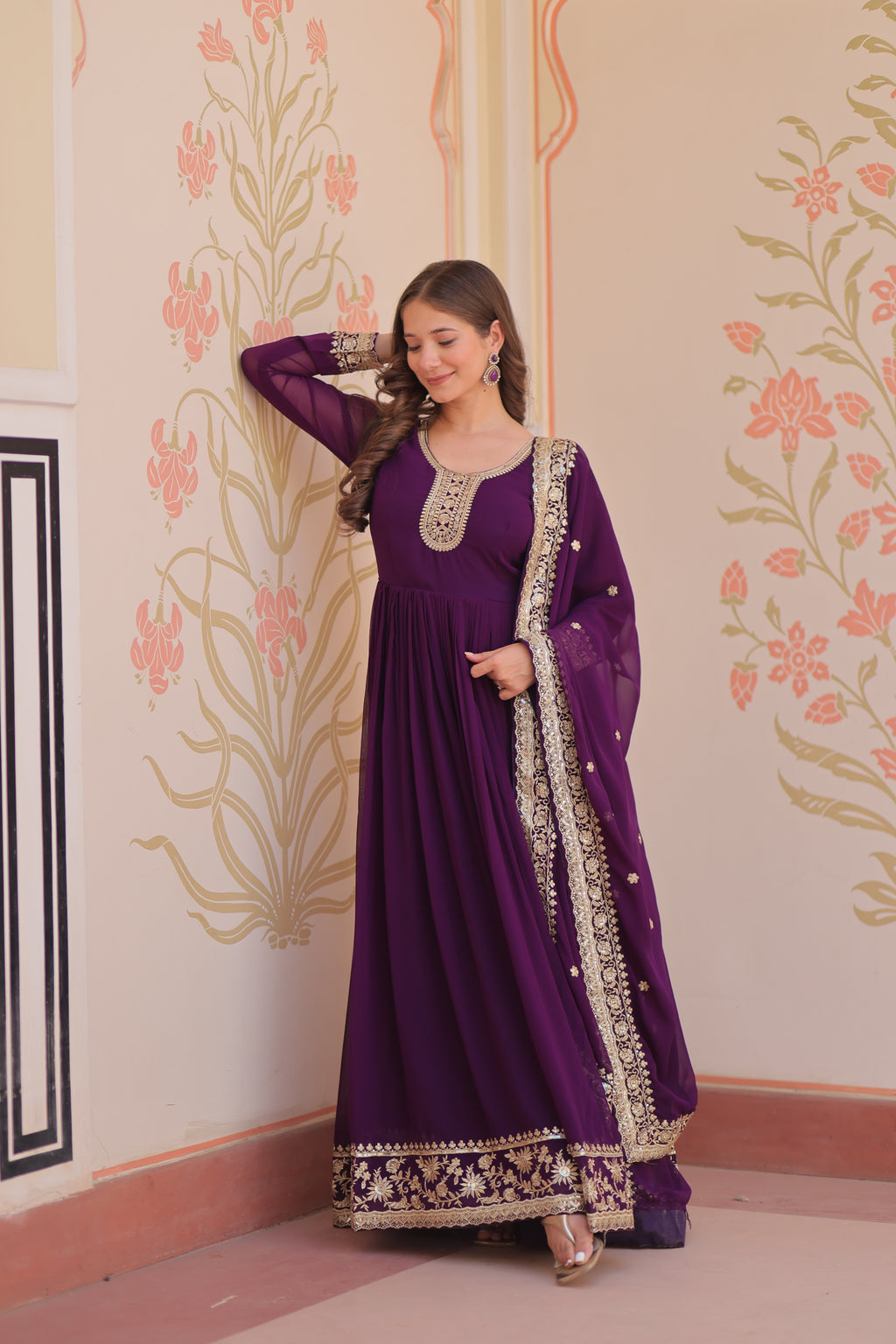 Woman in a purple dress with intricate designs standing against a decorated wall.