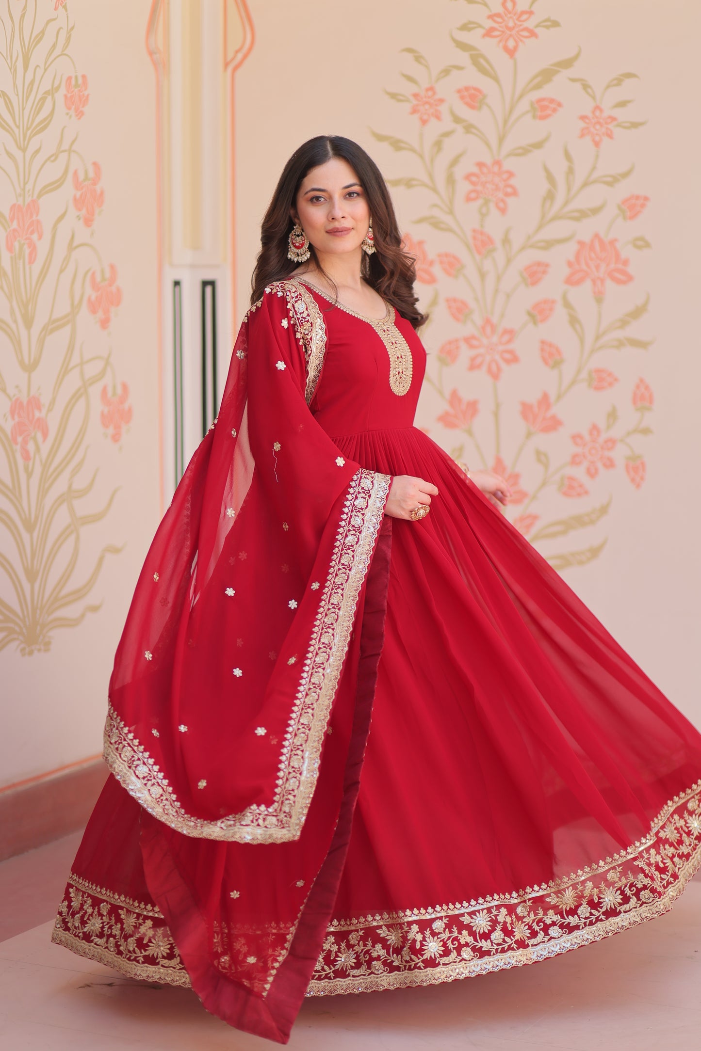 Woman wearing a red traditional outfit with gold embroidery against a floral wall.