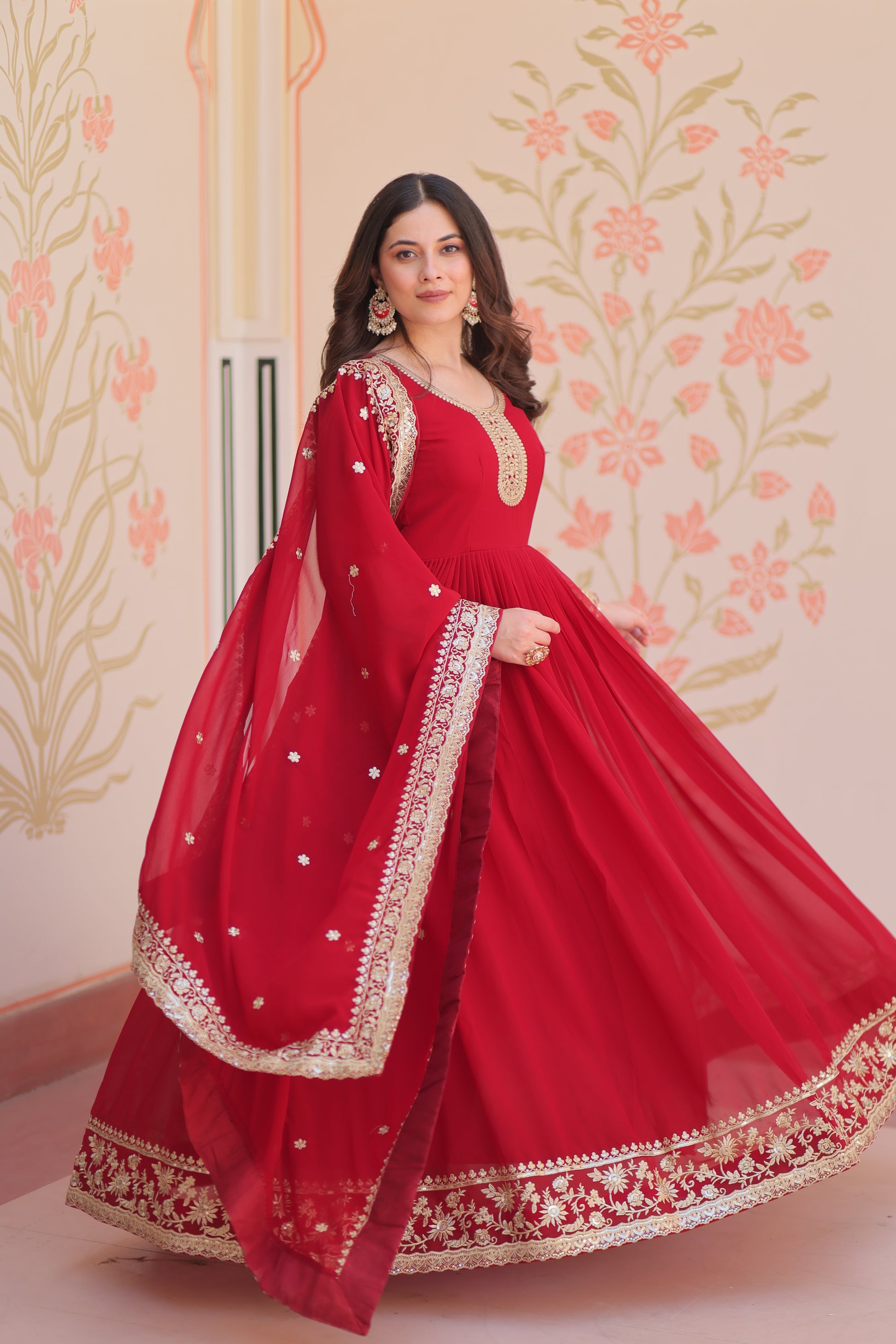 Woman wearing a red traditional outfit with gold embroidery against a floral wall.