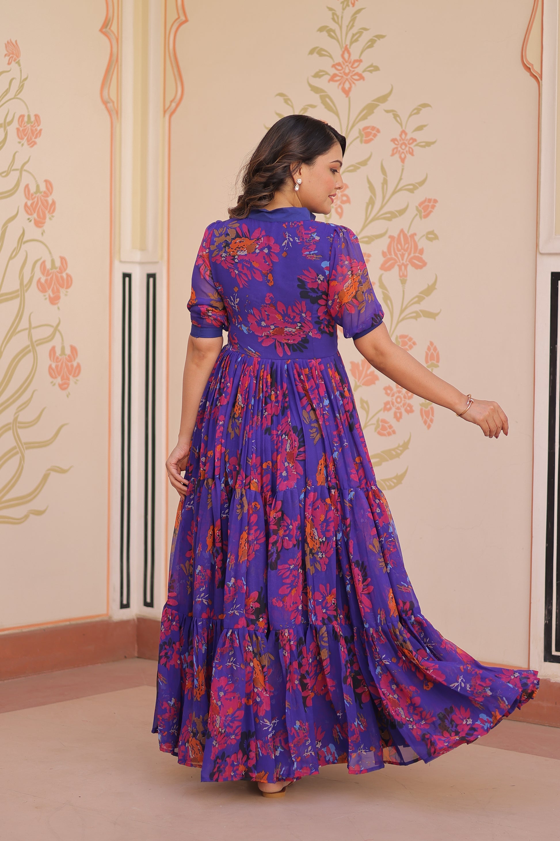 Woman wearing a colorful dress with floral patterns in a room with decorative wallpaper.