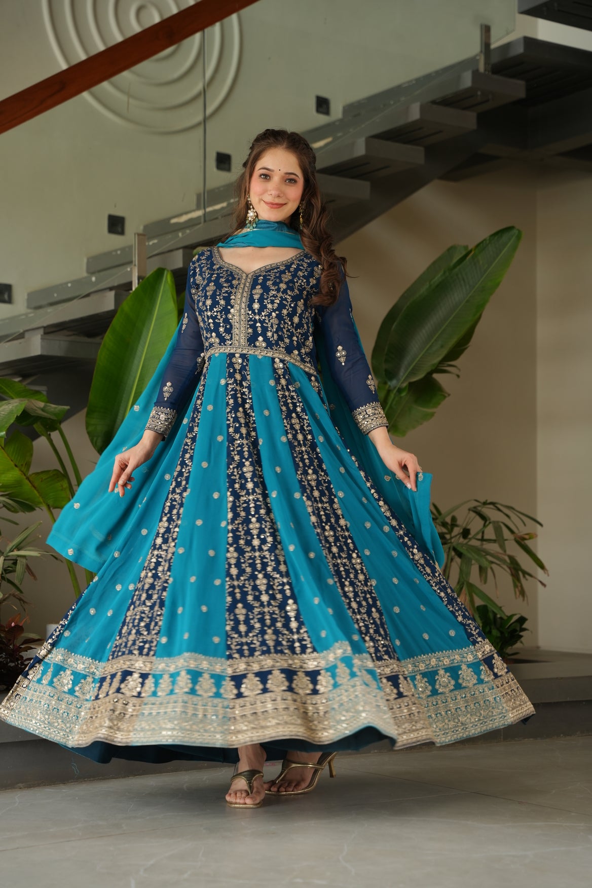 Woman in a blue and gold traditional dress standing indoors with plants in the background