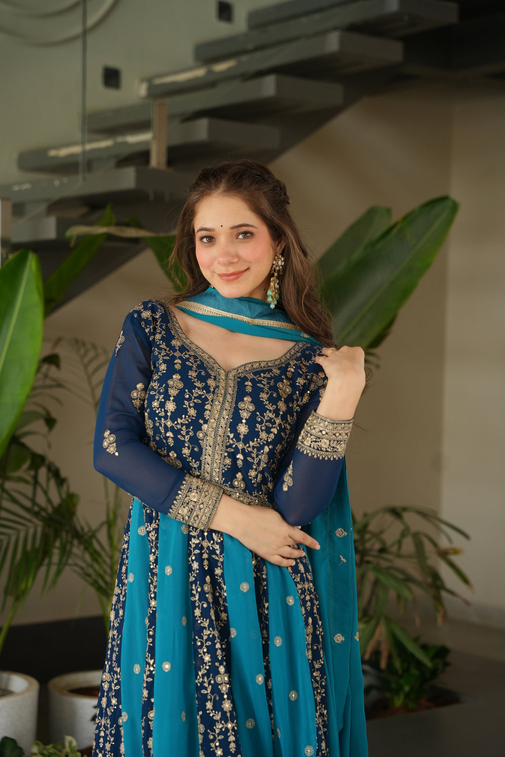 Woman in a blue and white traditional outfit standing indoors with plants in the background