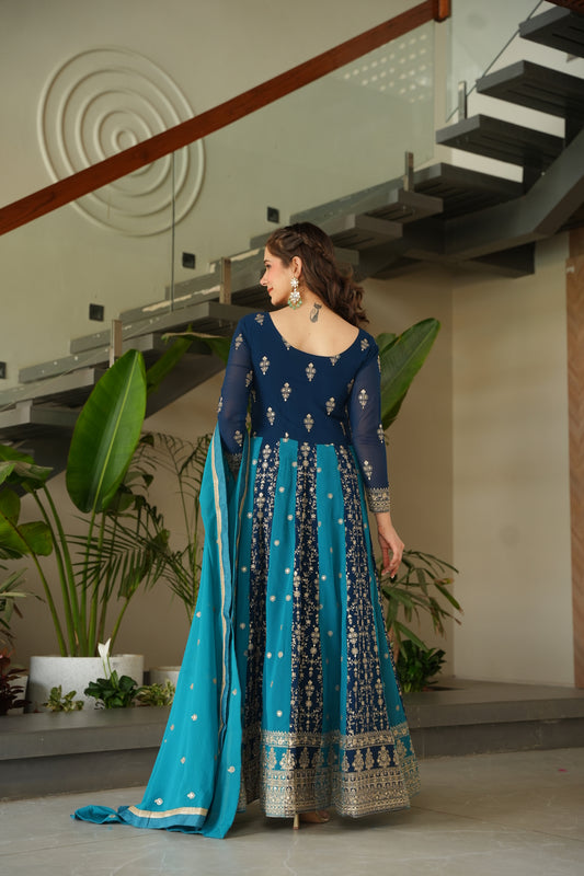 Woman in a blue and white traditional outfit standing indoors with plants in the background