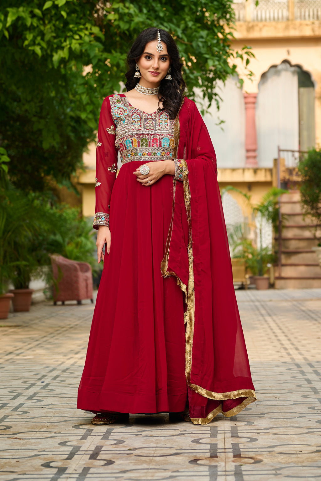 Woman in a red traditional outfit with intricate patterns standing outdoors.