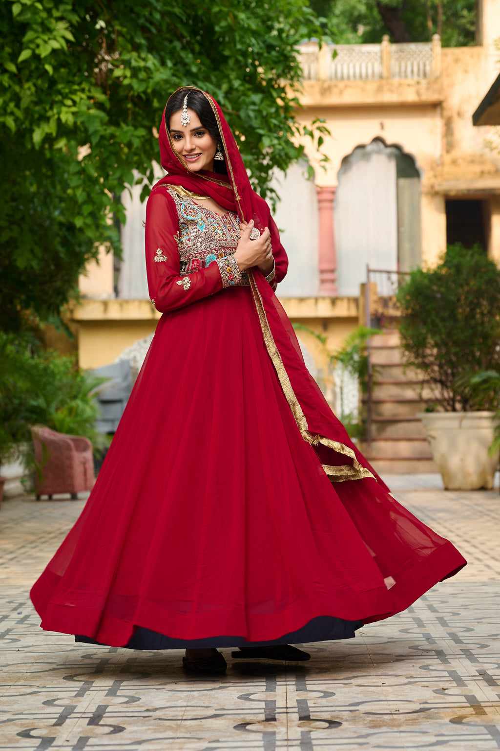 Woman in a red traditional outfit standing outdoors with greenery and architectural elements in the background