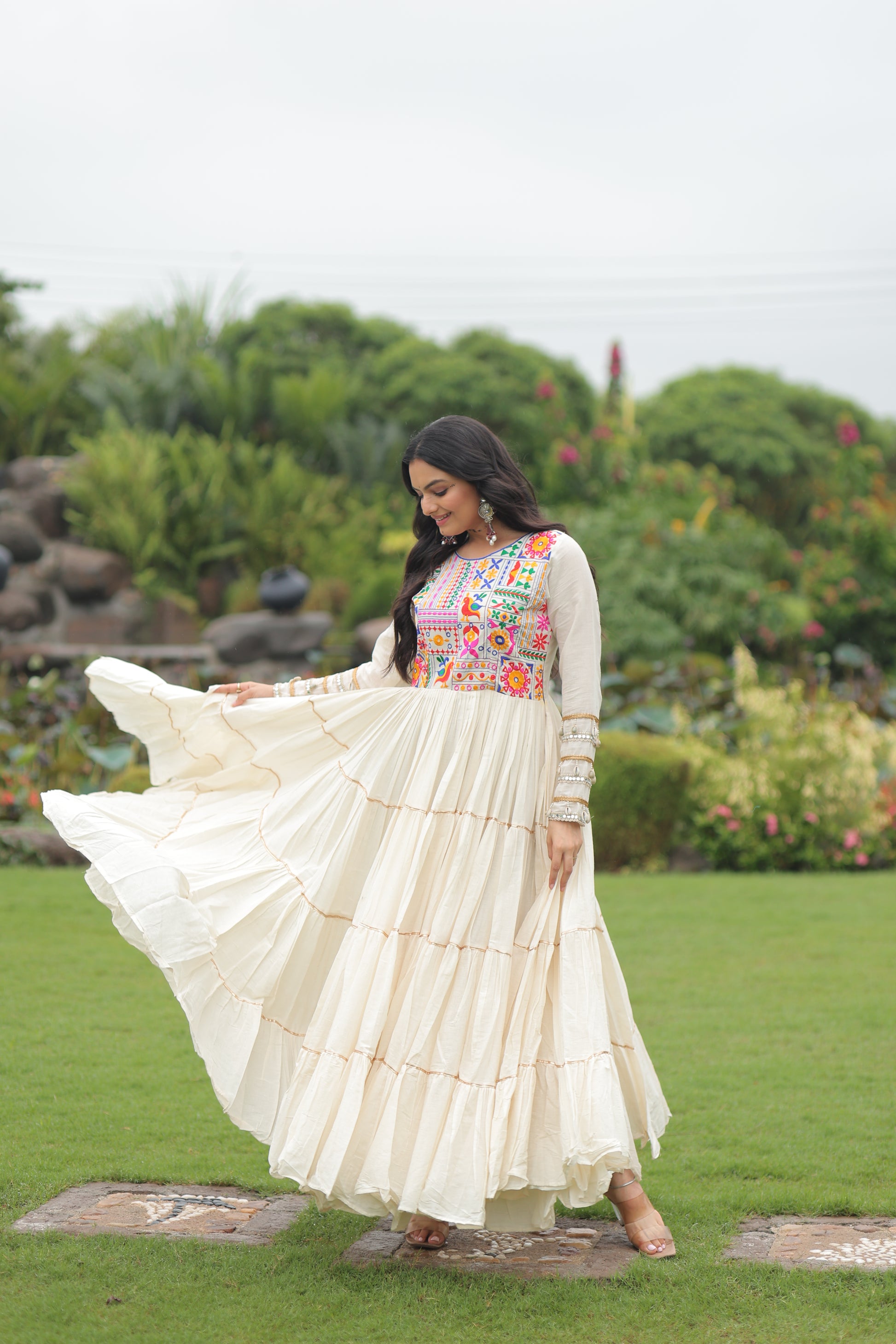 Woman in a white traditional outfit with floral patterns standing outdoors on grass.