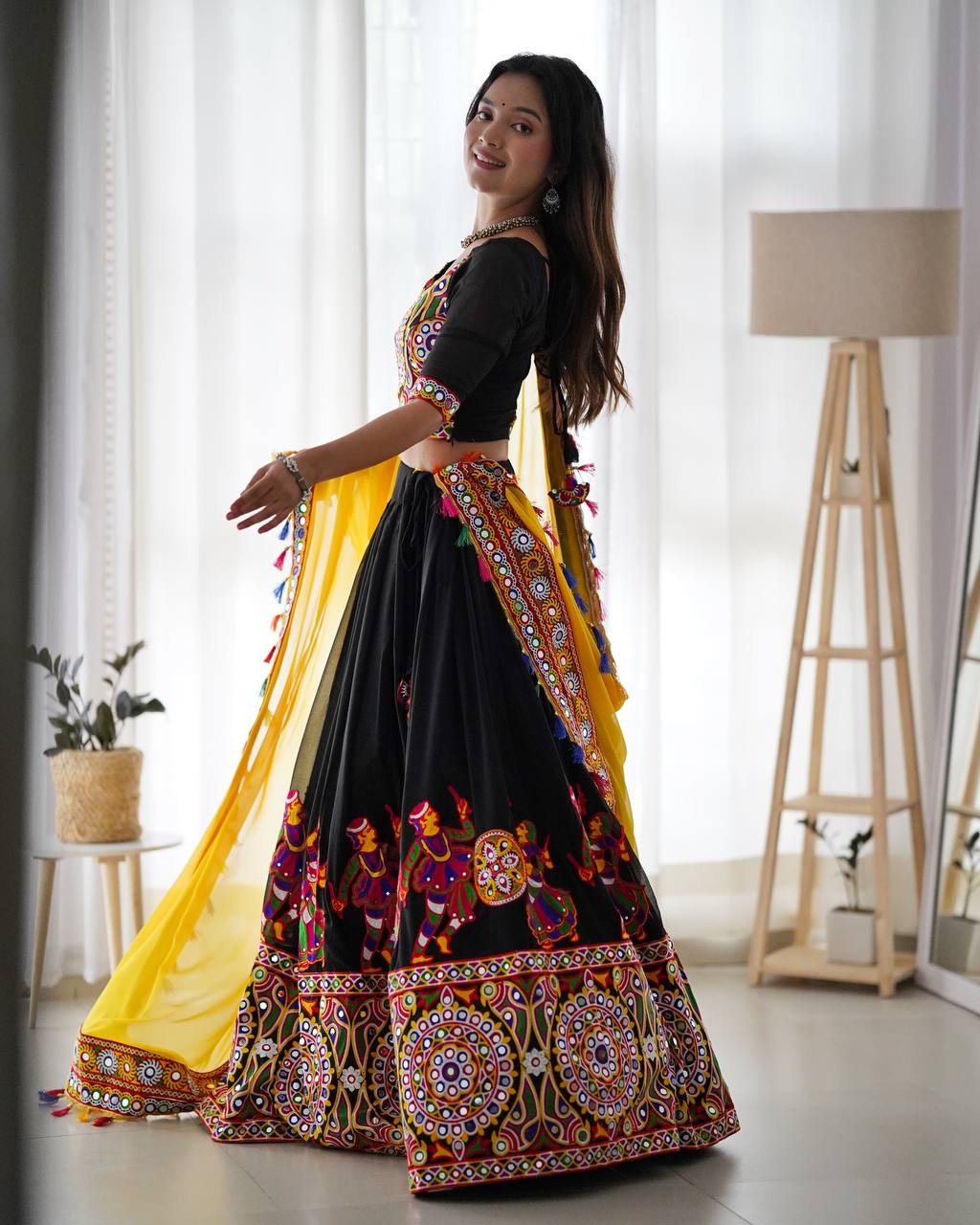 Woman in a traditional black and colorful outfit with a yellow dupatta indoors.