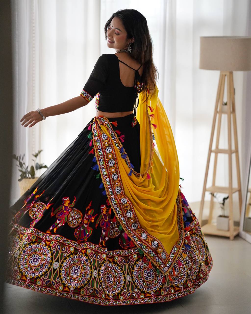 Woman in a traditional black and colorful lehenga with a yellow dupatta in a bright room.